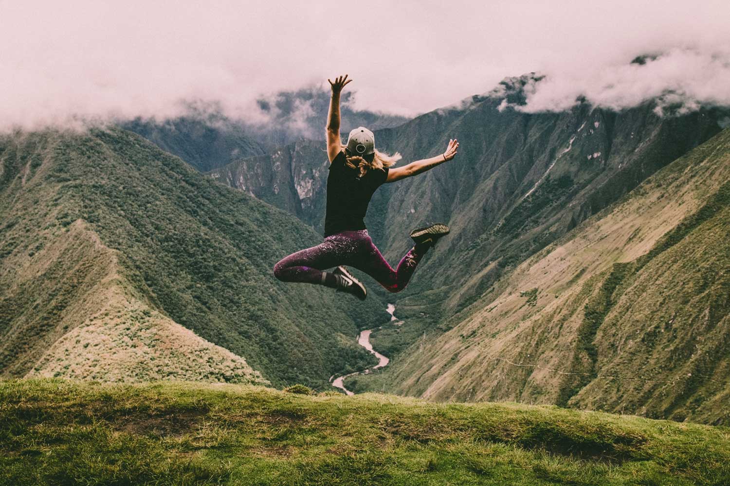 Junge Frau springt vor Freude in die Luft, im Hintergrund sind Berge und ein Fluss zu sehen